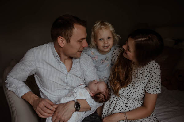 family with girl toddler and new baby boy son smile together as they sit on the bed
