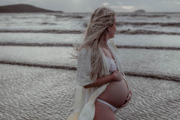 pregnant mother in cream robe holds baby bump and looks out into the ocean