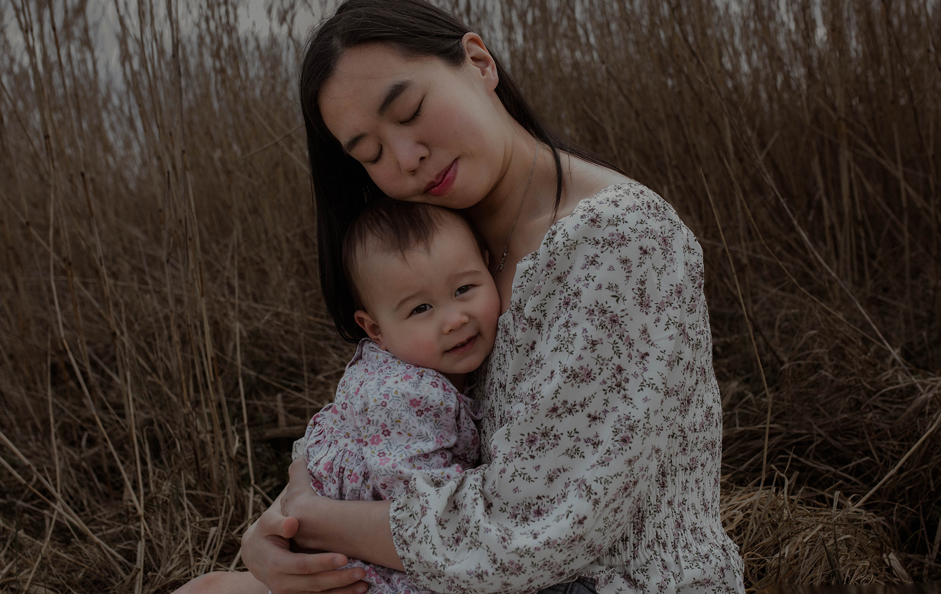 mother with dark hair in flowery shirt cuddles her little girl who is wearing a floral dress
