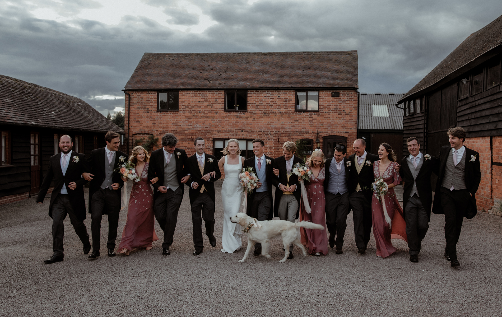 Group shot of wedding bridal party walking in a line smiling and laughing. Bride and groom stand in the middle as their dog runs along side them