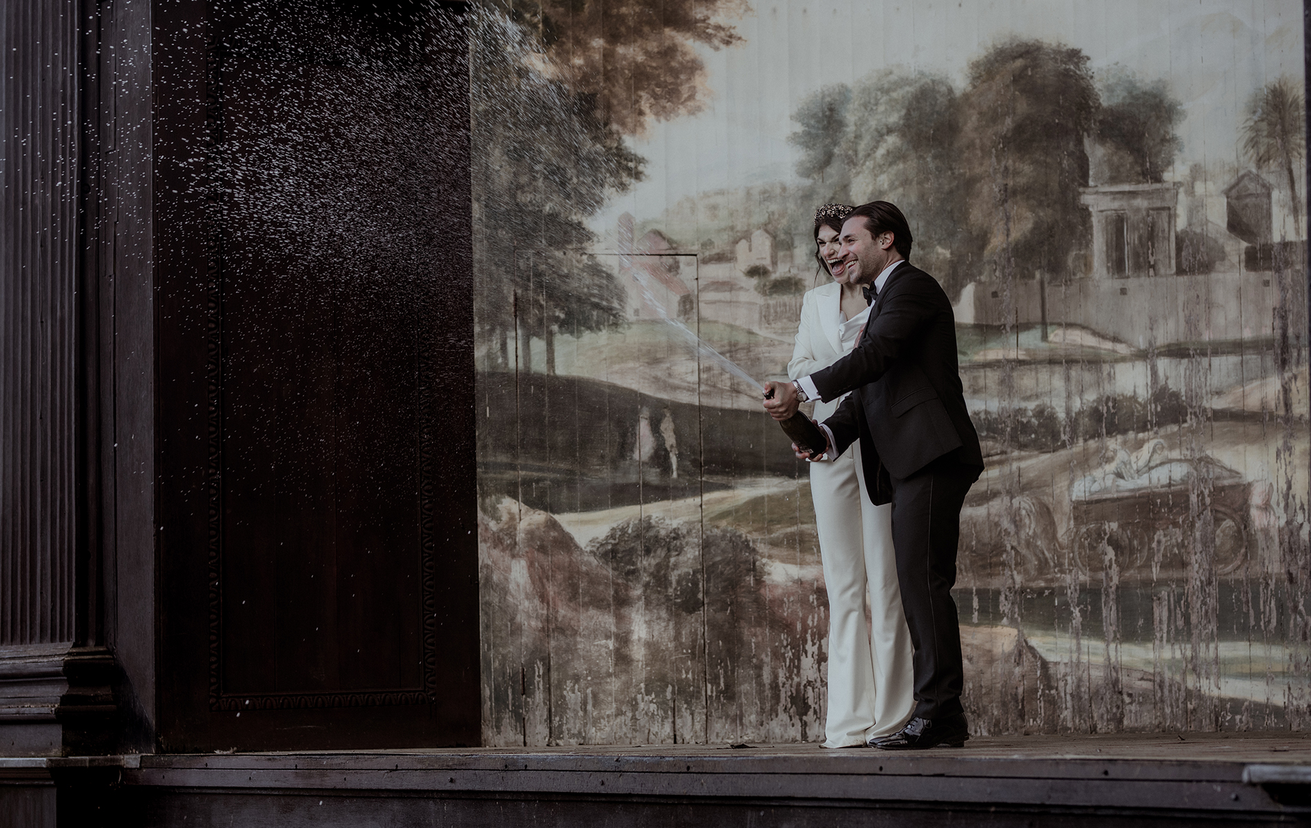 bride and groom in tuxedos spray champagne on a large mural stage