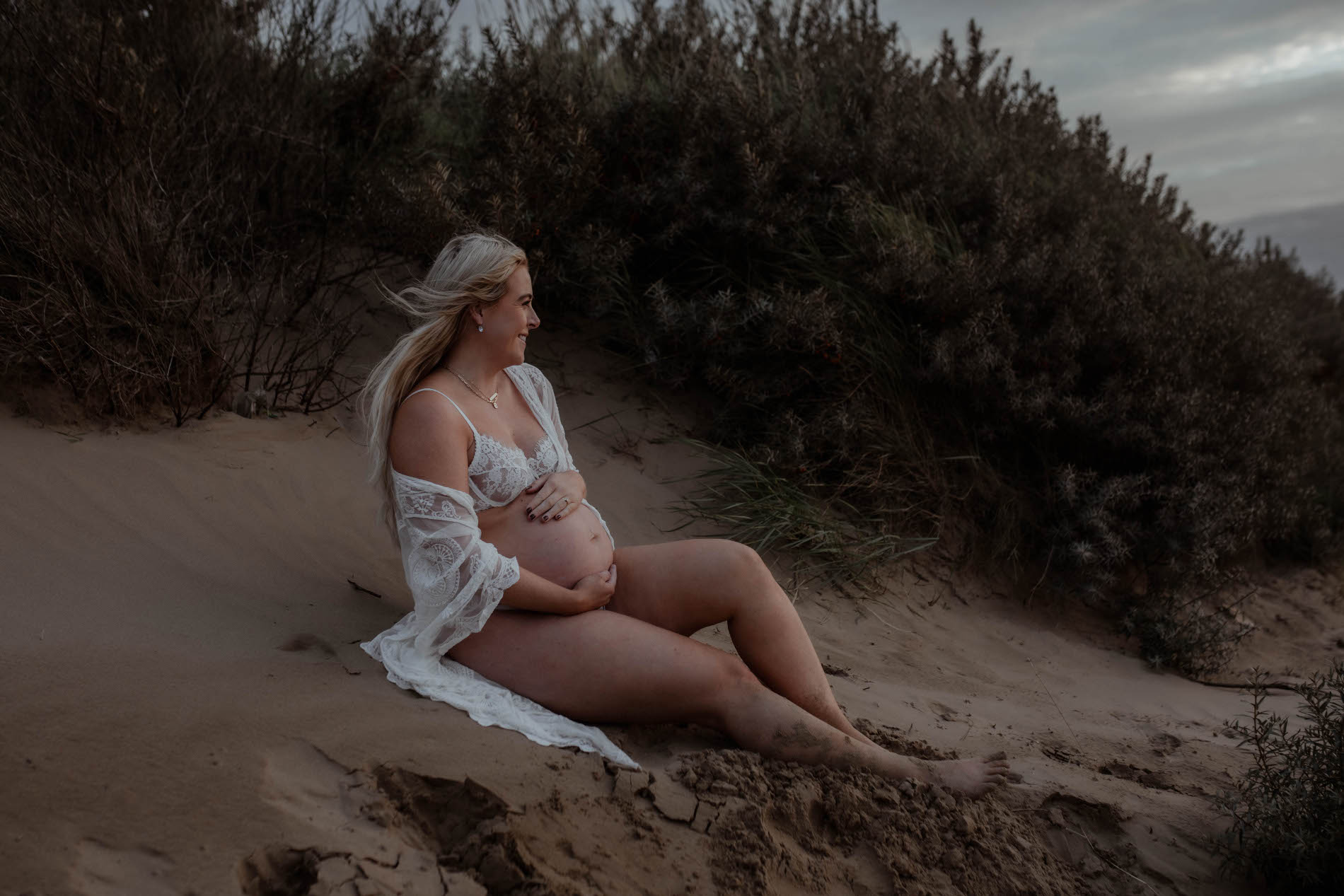 pregnant woman sits on bristol beach on a white robes and looks off into the distance