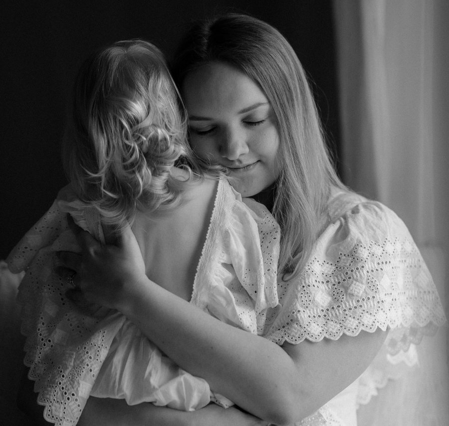 black and white image where mother and young daughter hug each other, both are wearing white frilled dresses